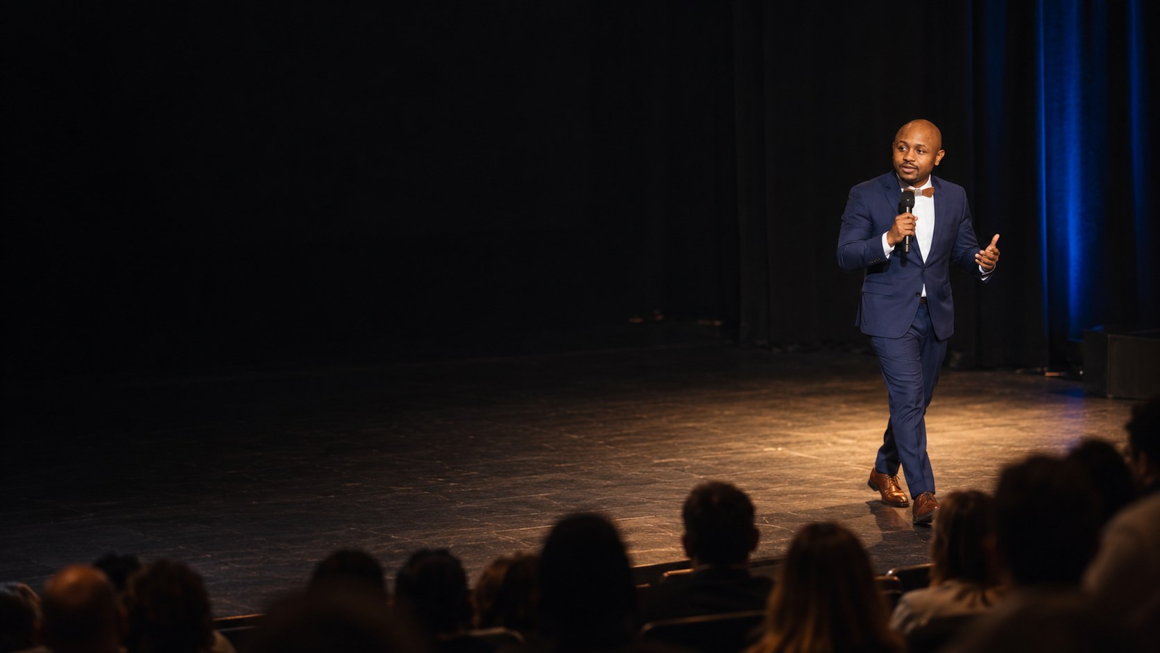 Ryan Perez walking a dark stage with a microphone, audience in silhouette